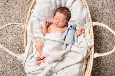 Photo of Cute newborn baby sleeping with toy bear on white blanket in wicker crib, top view