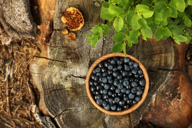 Photo of Wooden bowl of delicious bilberries and green leaves on stump, flat lay. Space for text