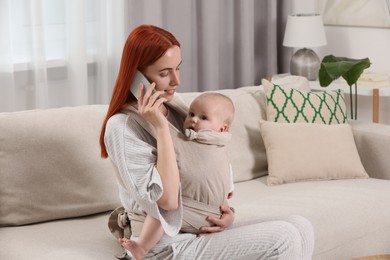 Photo of Mother talking on smartphone while holding her child in sling (baby carrier) at home