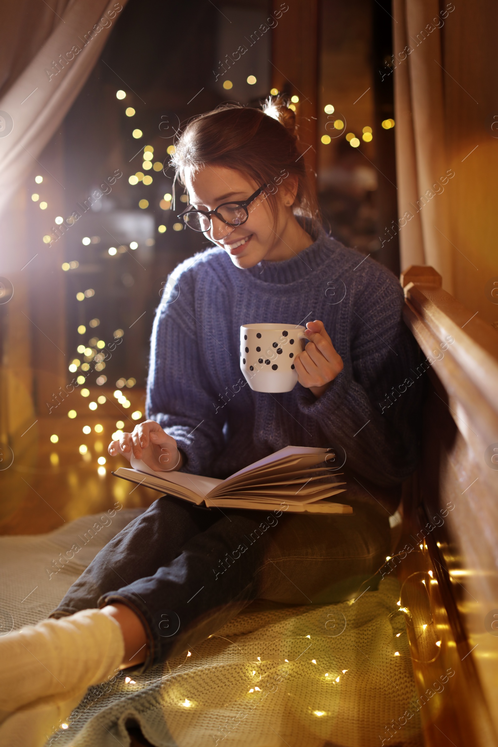 Photo of Woman with cup of hot beverage reading book at home in winter evening