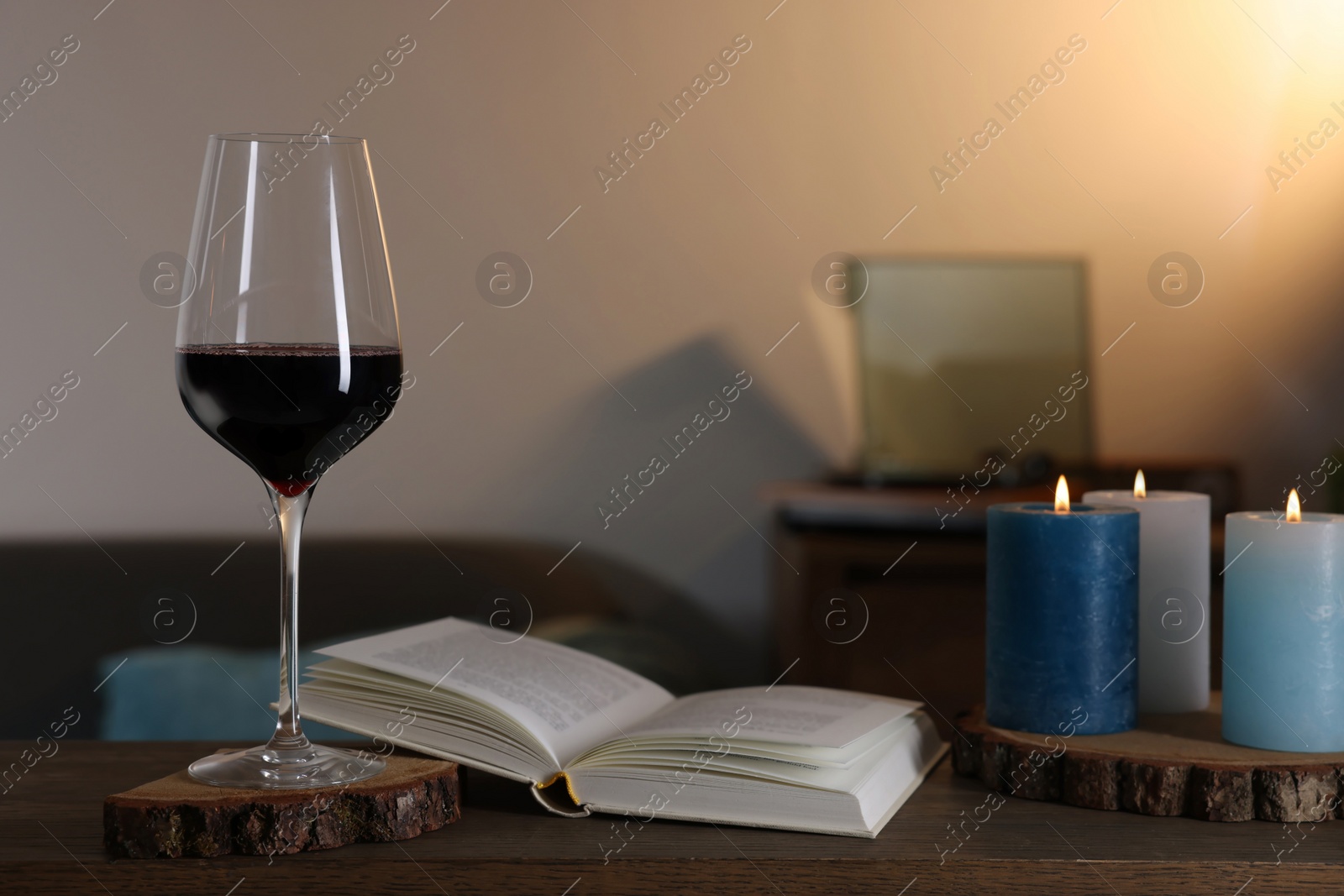 Photo of Glass of red wine, open book and burning candles on wooden table in room. Relax at home