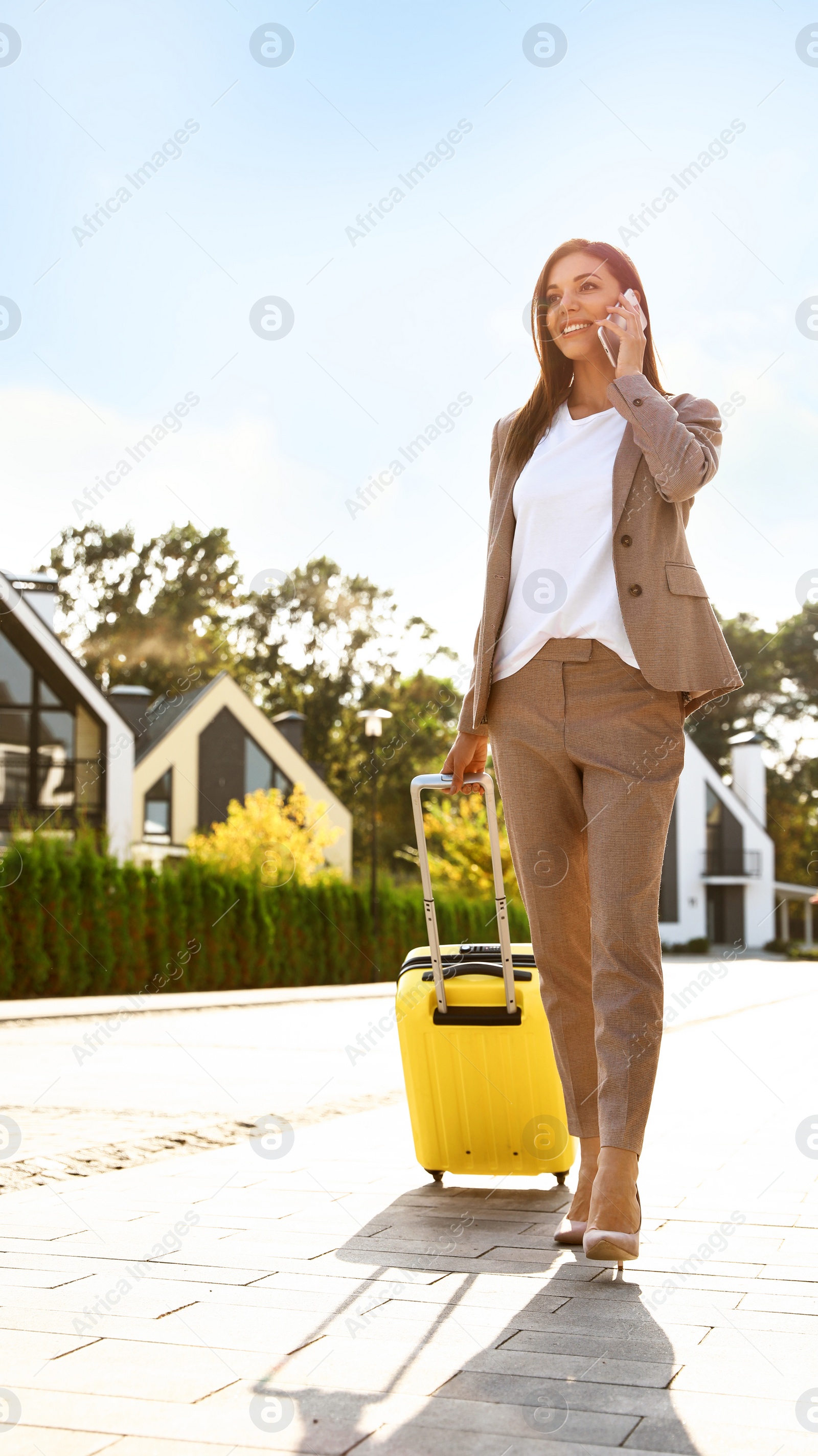 Photo of Young businesswoman with suitcase talking on mobile phone outdoors. Moving day