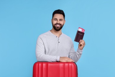 Smiling man with passport, tickets and suitcase on light blue background