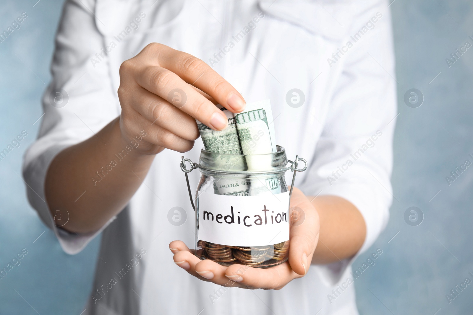 Photo of Woman holding glass jar with money and tag MEDICATION on light blue background, closeup