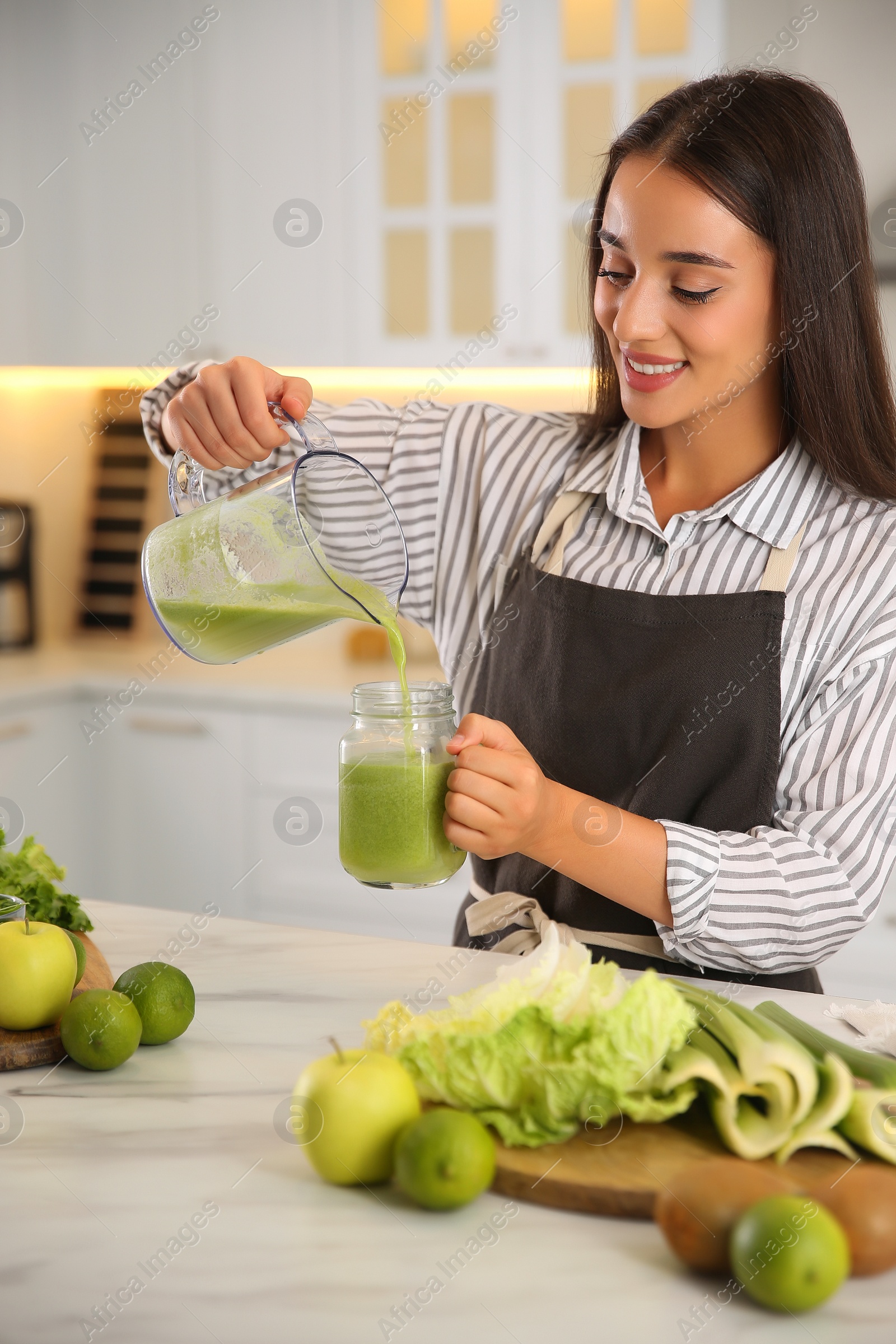 Photo of Young woman pouring fresh green juice into mason jar at table in kitchen