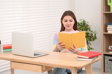 Photo of Cute girl reading book near laptop at desk in room. Home workplace