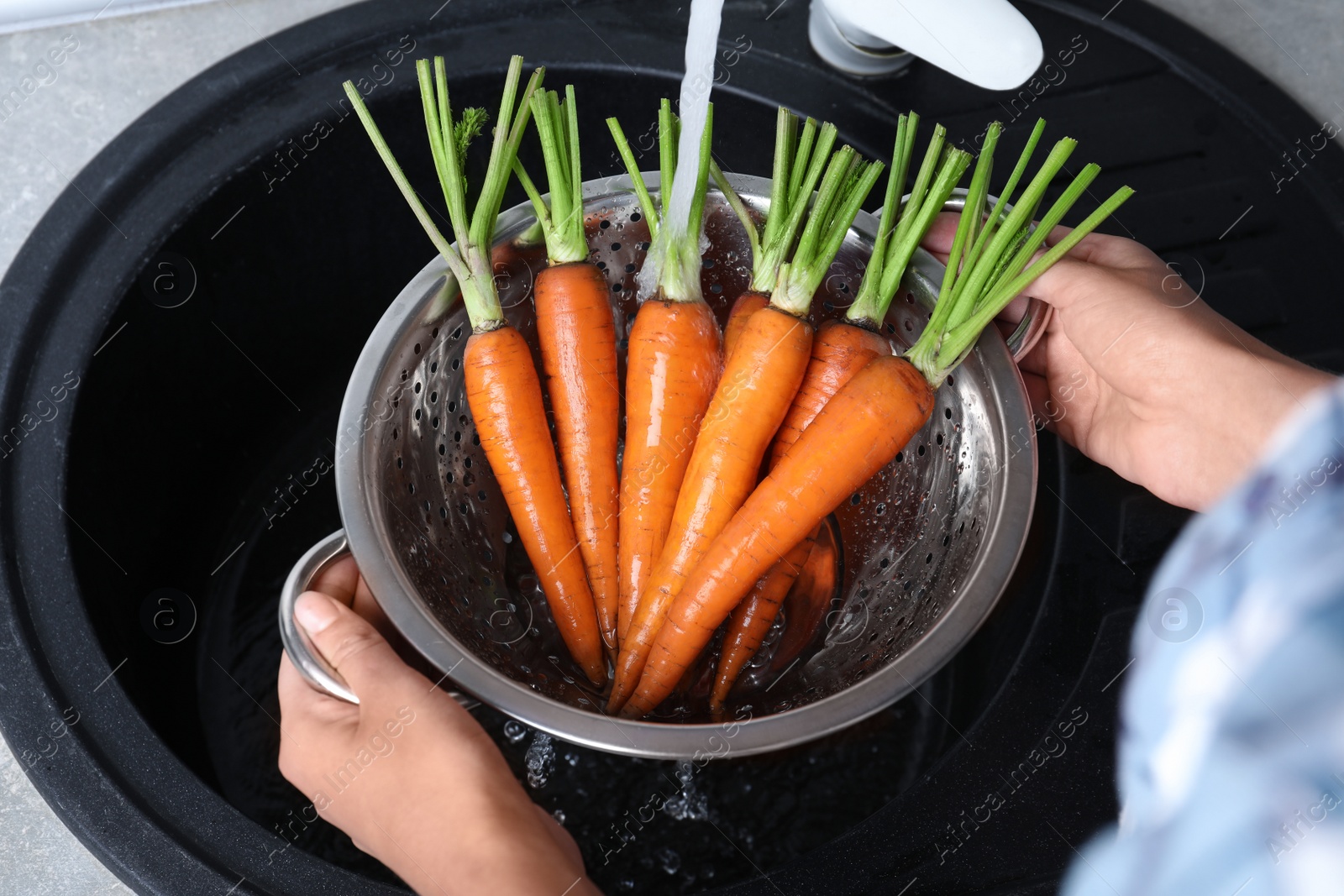 Photo of Woman washing ripe carrots in colander with running water over sink, closeup