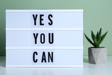 Photo of Lightbox with phrase Yes You Can and potted houseplant on table against light green background. Motivational quote