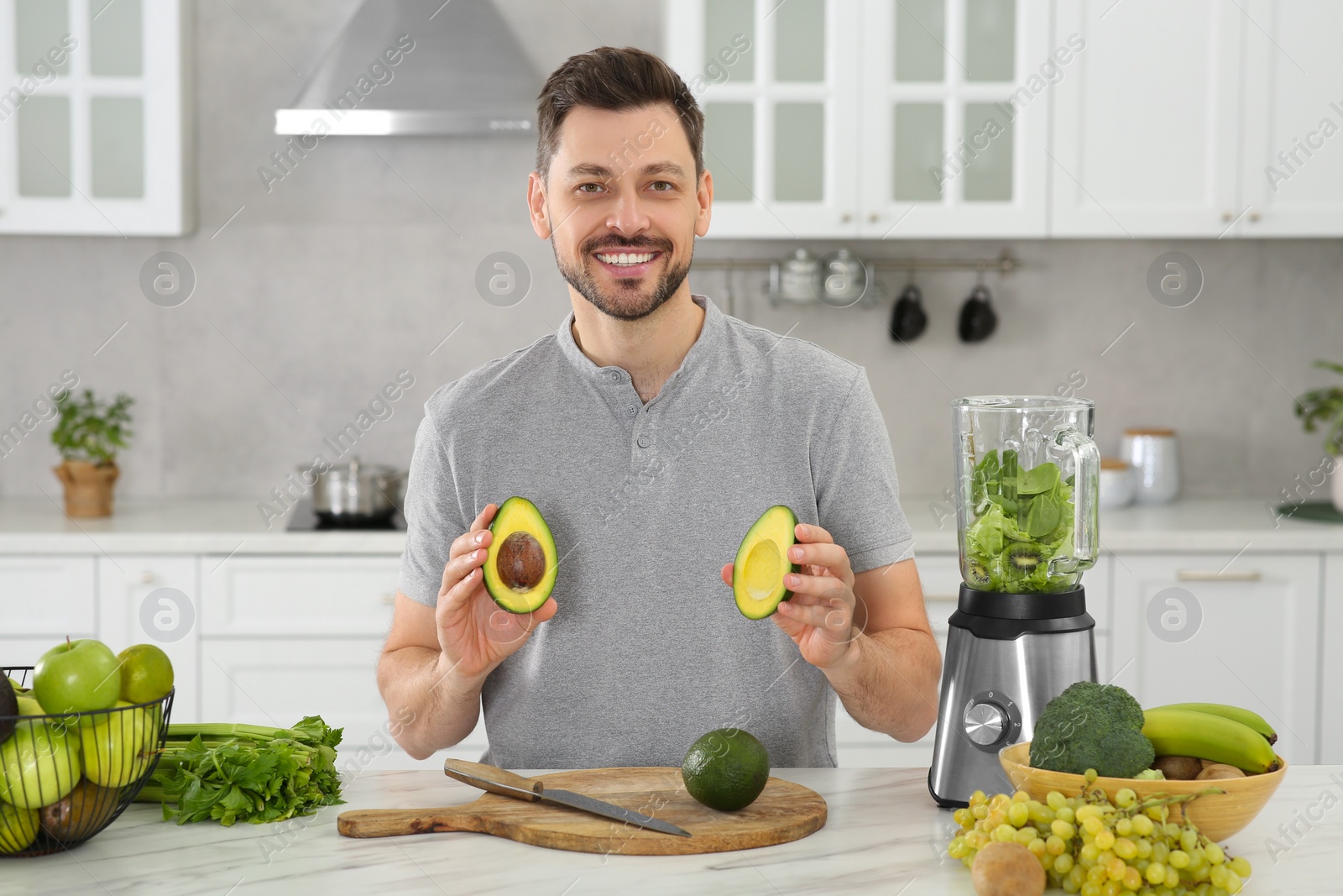 Photo of Man holding halves of avocado for delicious smoothie at white marble table in kitchen