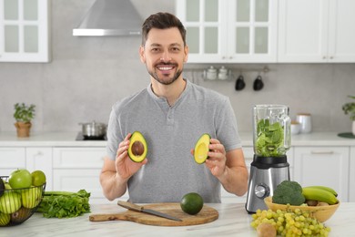 Photo of Man holding halves of avocado for delicious smoothie at white marble table in kitchen