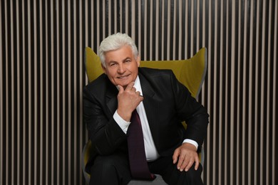 Happy mature businessman sitting in armchair near wood slat wall