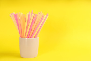 Colorful plastic drinking straws in holder on yellow background, space for text
