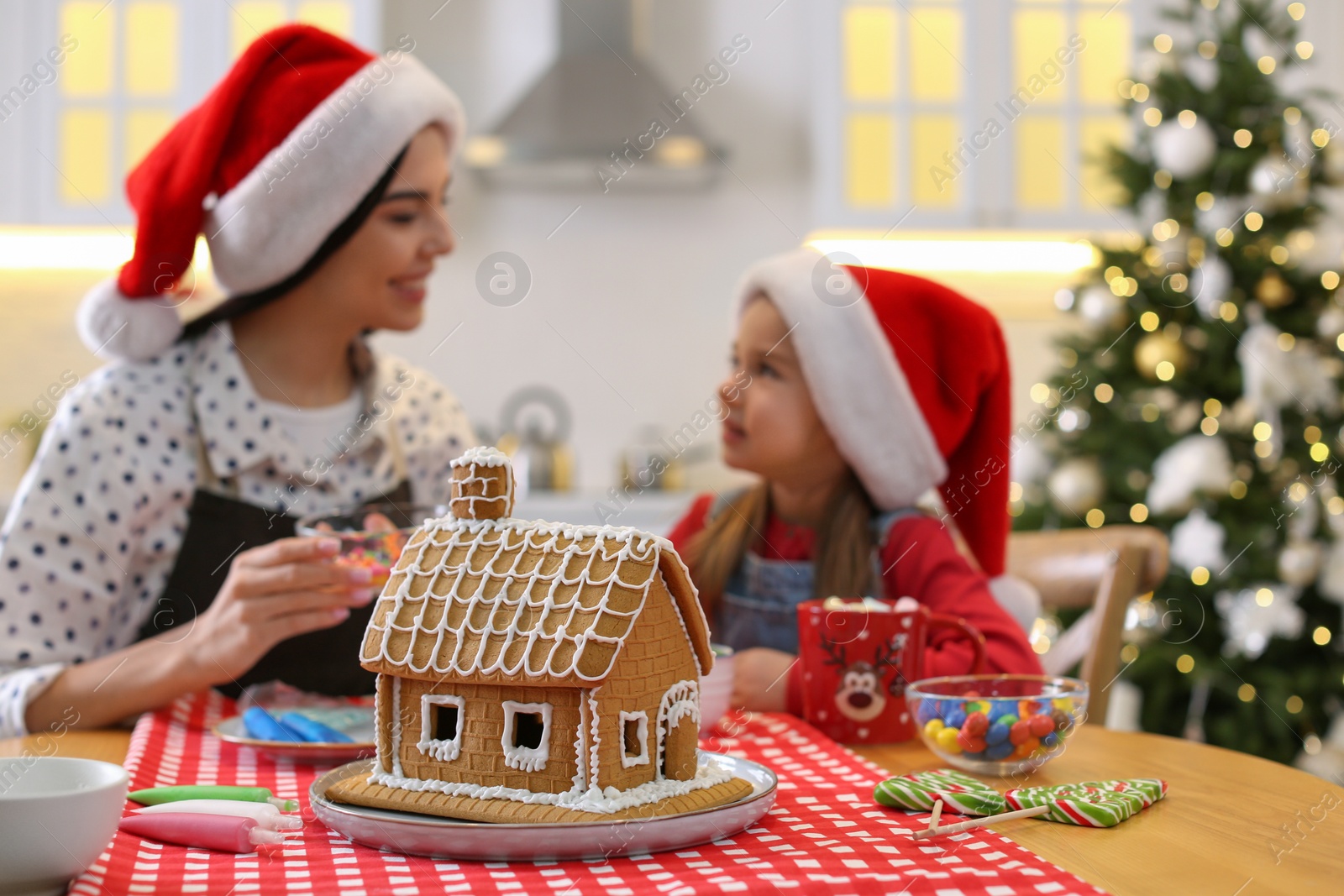 Photo of Mother and daughter in Santa hats cooking together, focus on gingerbread house