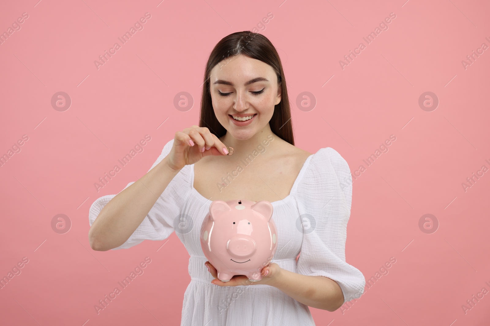 Photo of Happy woman putting coin into piggy bank on pink background