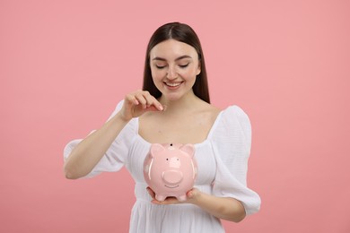 Happy woman putting coin into piggy bank on pink background