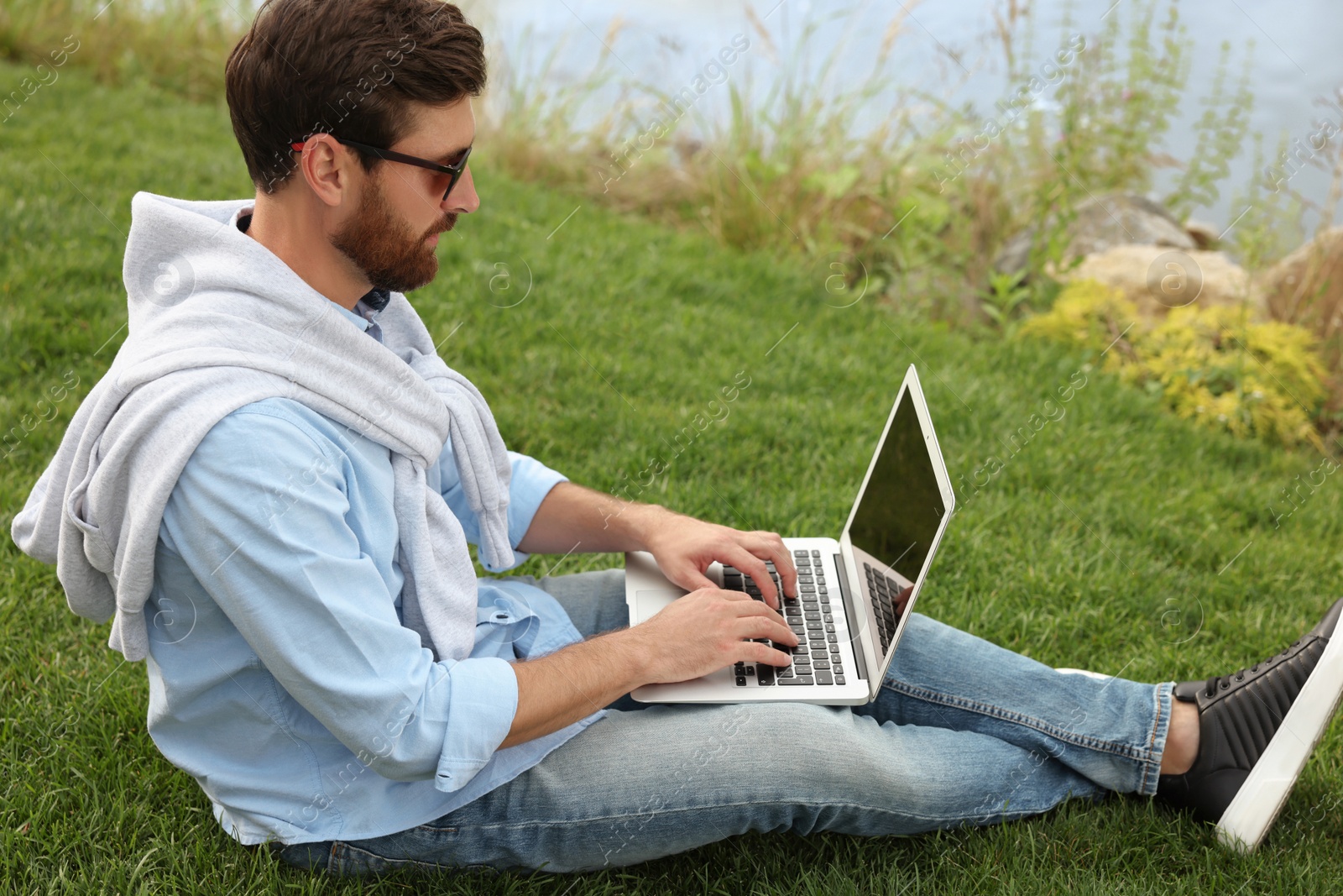 Photo of Man in sunglasses with laptop on green grass near lake