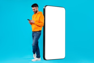 Image of Man with mobile phone standing near huge device with empty screen on light blue background. Mockup for design