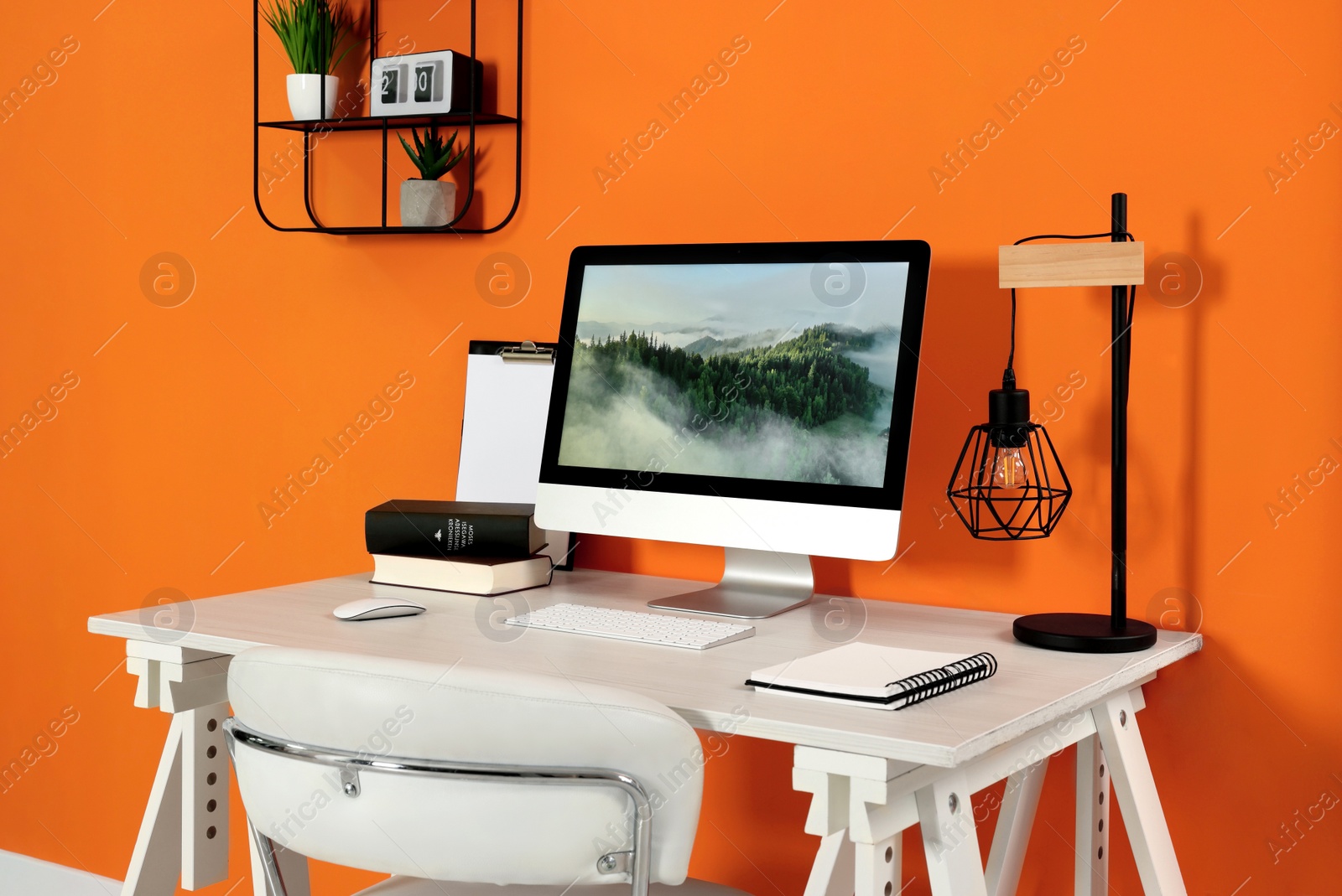 Photo of Modern computer, books, lamp and notebook on wooden desk near orange wall. Home office