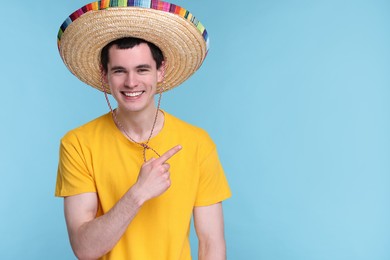 Photo of Young man in Mexican sombrero hat pointing at something on light blue background. Space for text