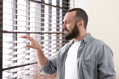 Portrait of handsome mature man looking through window blinds indoors