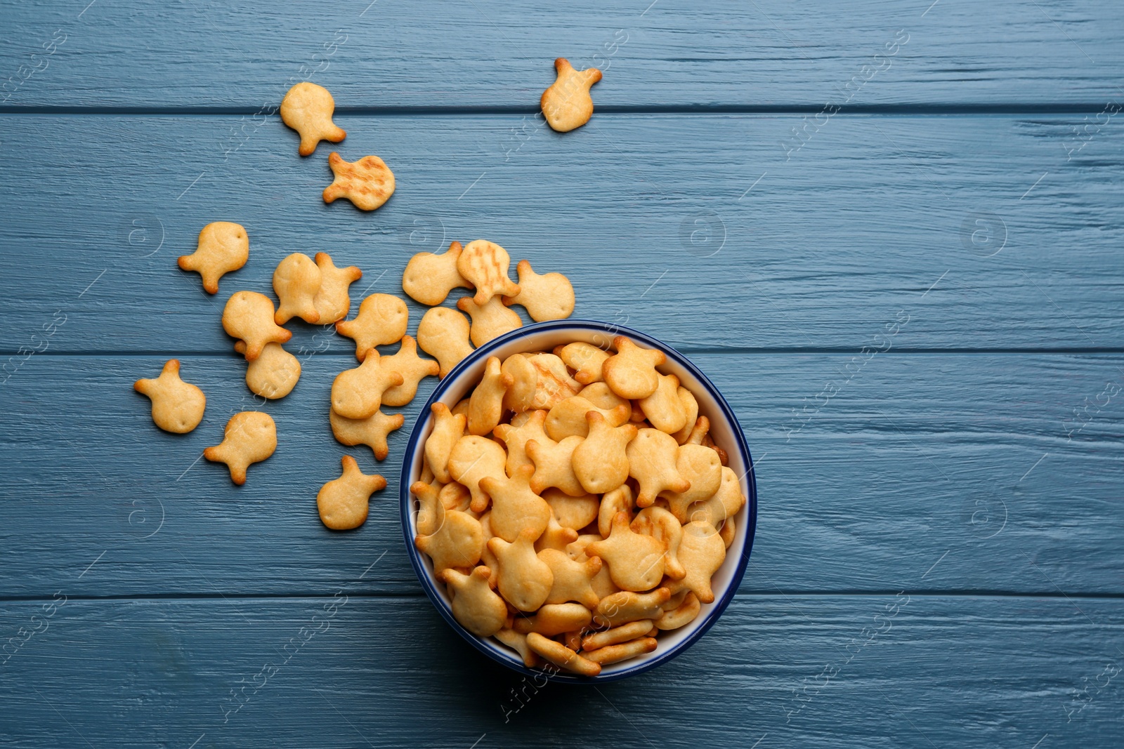 Photo of Delicious goldfish crackers in bowl on blue wooden table, flat lay