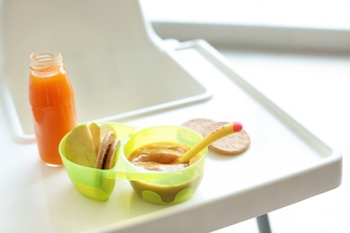 Bowls with delicious baby food and bottle of juice on highchair indoors