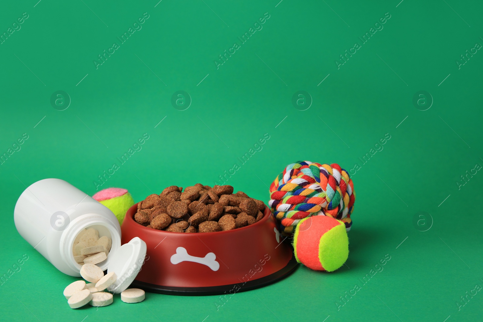 Photo of Bowl with dry pet food, bottle of vitamins and toys on green background. Space for text