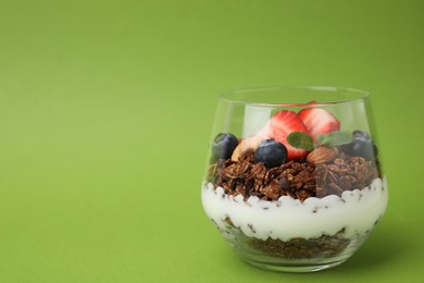 Photo of Tasty granola with berries, nuts and yogurt in glass on green background, space for text