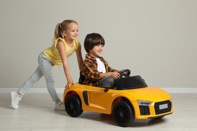 Cute girl pushing children's electric toy car with little boy near grey wall indoors