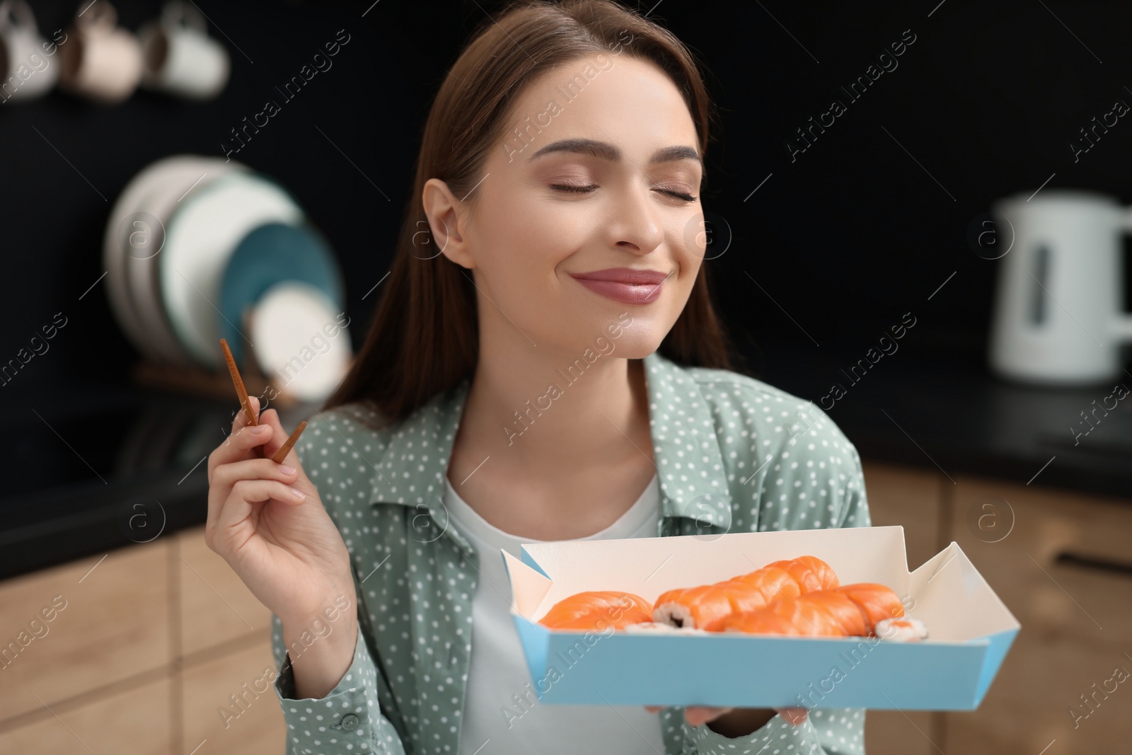 Photo of Beautiful young woman eating sushi rolls with chopsticks in kitchen