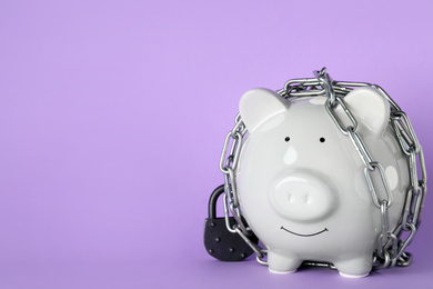Photo of Piggy bank  with steel chain and padlock on lilac background, space for text. Money safety concept