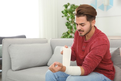 Young man applying bandage on injured arm at home, space for text. First aid