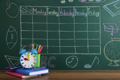 Photo of Alarm clock and stationery on wooden table near green chalkboard with drawn school timetable