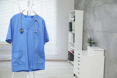 Blue medical uniform and stethoscope hanging on rack in clinic. Space for text