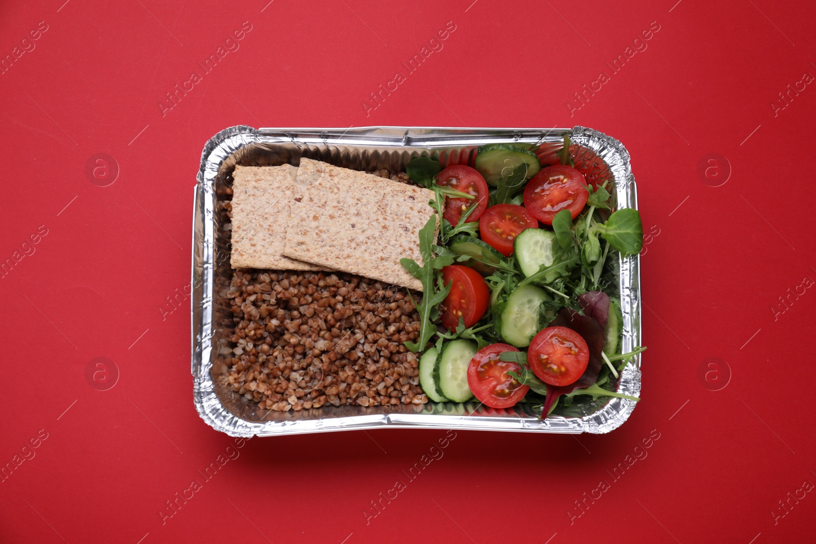 Photo of Container with buckwheat, fresh salad and crispbreads on red background, top view