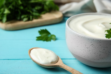 Photo of Bowl of fresh sour cream with parsley and spoon on light blue wooden table