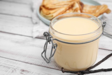 Photo of Tasty condensed milk in jar, crepes and vanilla pods on white wooden table, closeup. Space for text