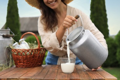 Photo of Smiling woman pouring fresh milk from can into glass at wooden table outdoors, closeup