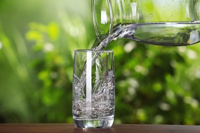 Photo of Pouring water from jug into glass on wooden table outdoors, closeup