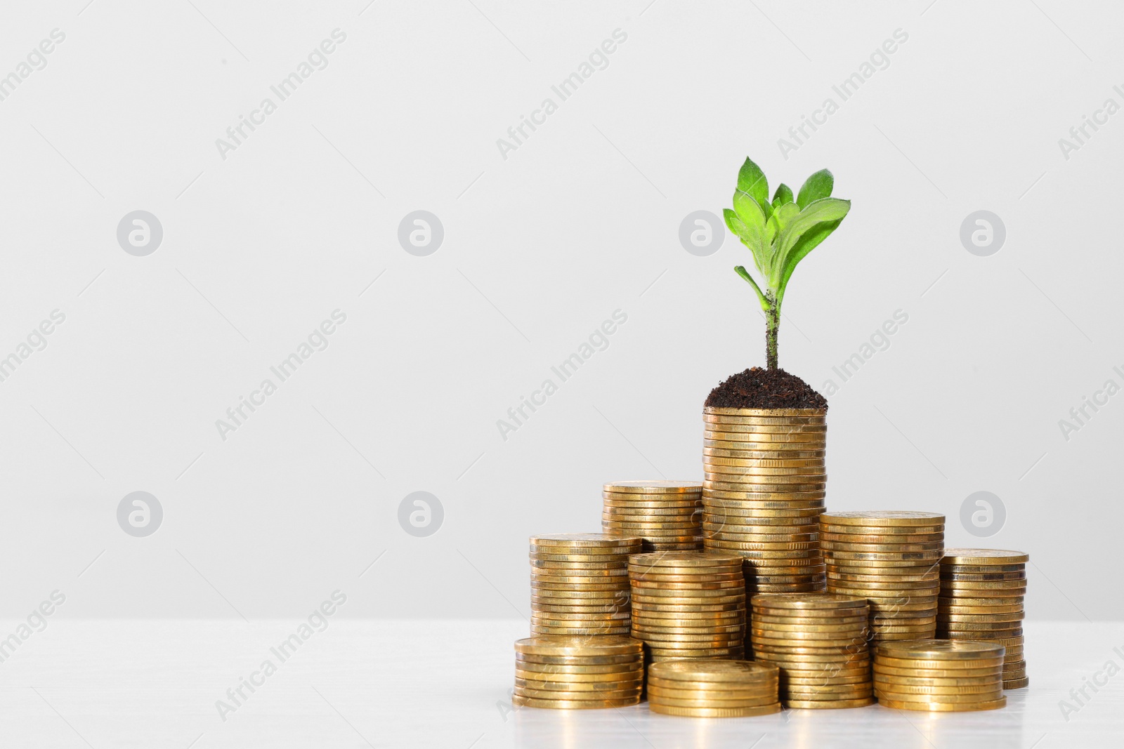 Photo of Stacks of coins with green sprout on white table against light grey background, space for text. Investment concept