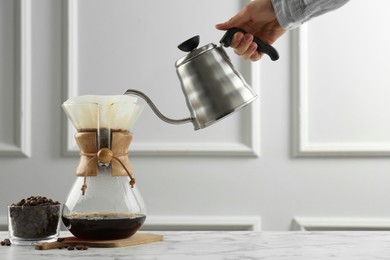 Man pouring hot water from kettle into glass chemex coffeemaker on white marble table, closeup