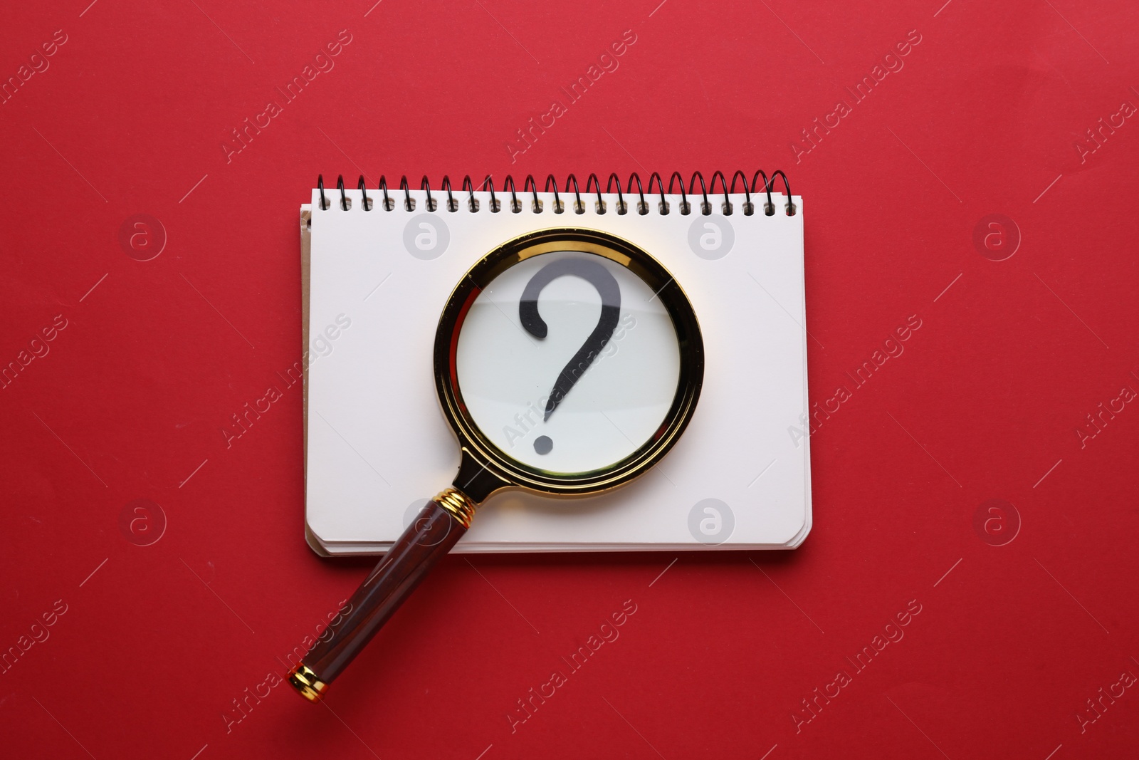 Photo of Magnifying glass over notebook with question mark on red background, top view