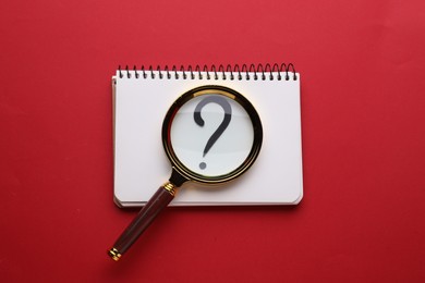 Magnifying glass over notebook with question mark on red background, top view