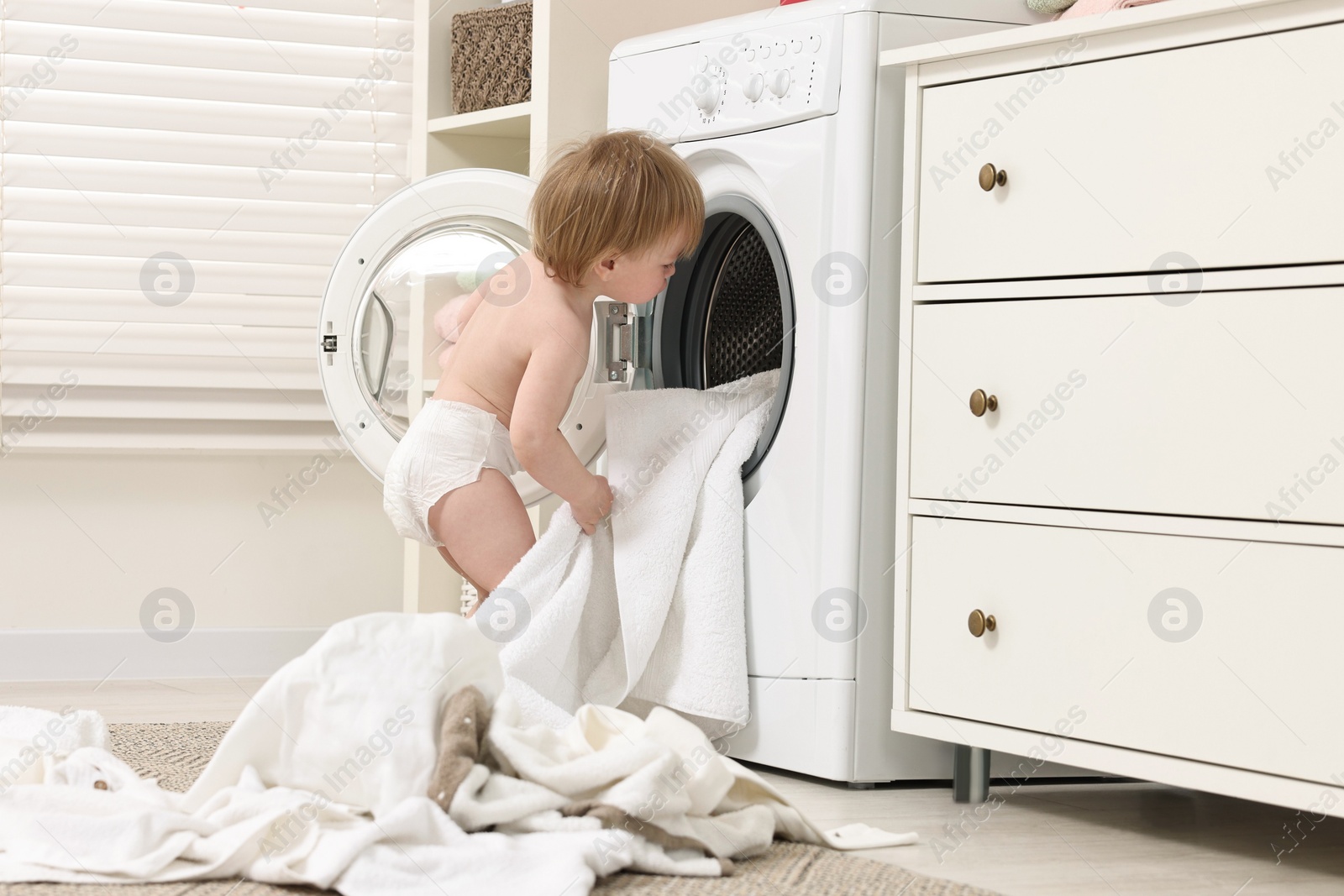 Photo of Little girl pulling baby clothes out of washing machine indoors