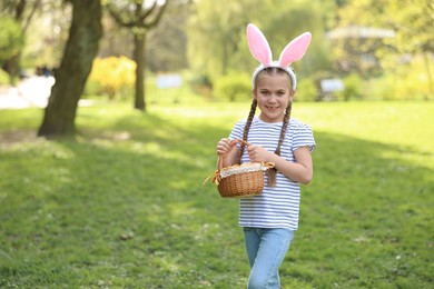 Easter celebration. Cute little girl in bunny ears holding wicker basket outdoors, space for text