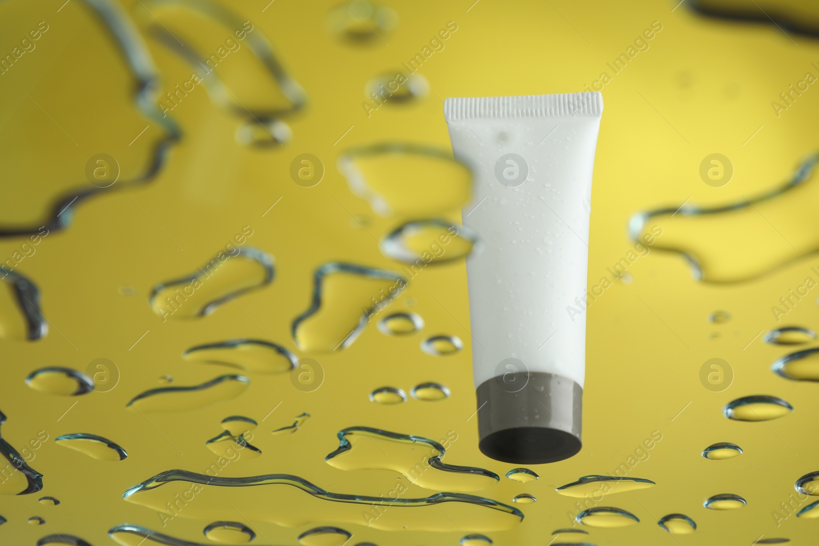 Photo of Moisturizing cream in tube on glass with water drops against yellow background, low angle view. Space for text