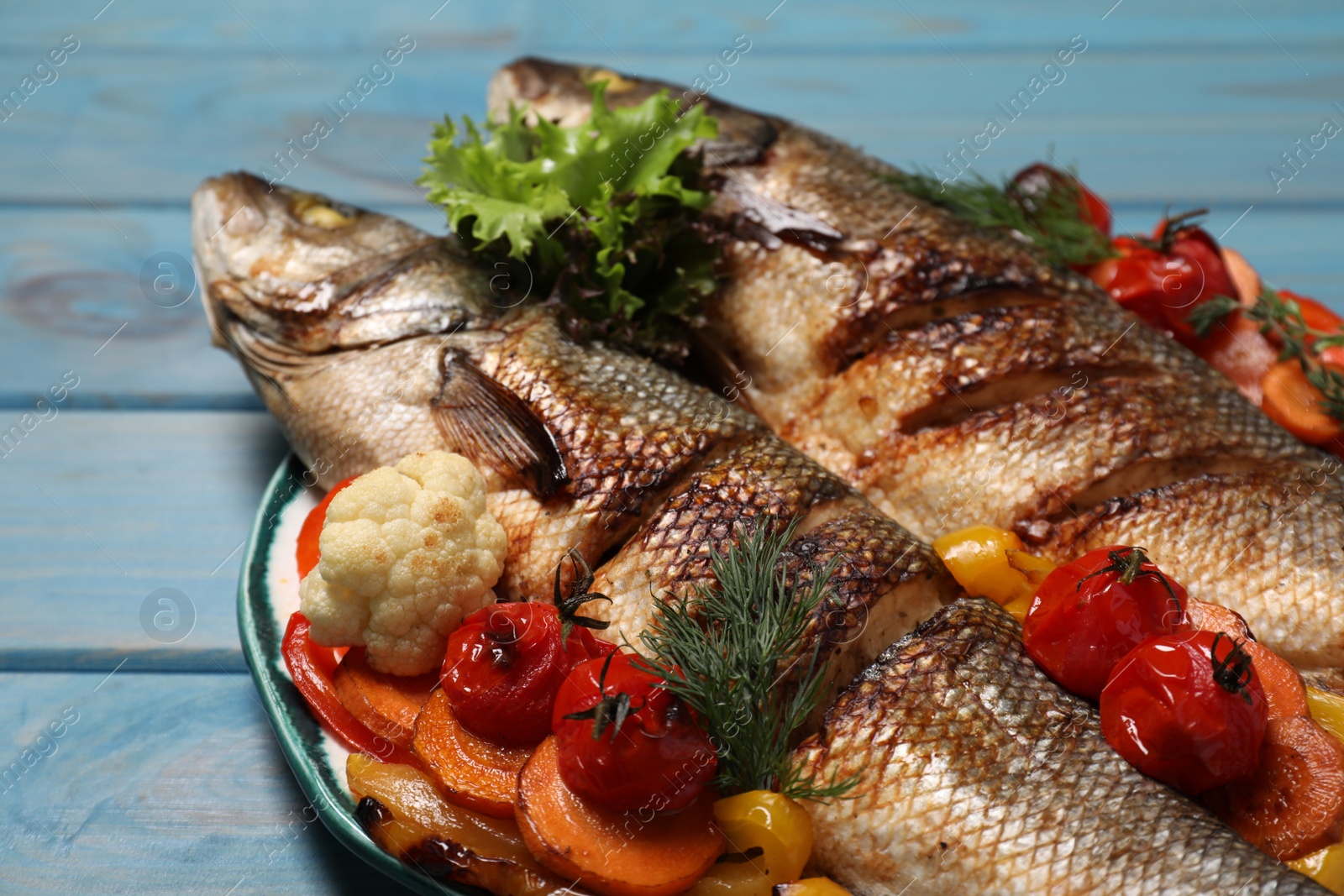 Photo of Plate with delicious roasted sea bass fish and vegetables on light blue wooden table, closeup