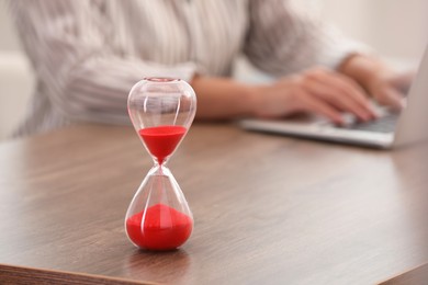 Photo of Hourglass with red flowing sand on table. Woman using laptop indoors, closeup