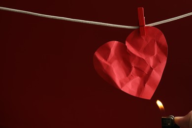 Photo of Lighting red paper heart with lighter on rope against burgundy background, space for text. Broken heart