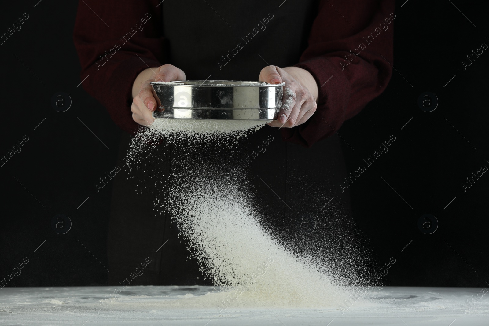 Photo of Woman sieving flour at table against black background, closeup